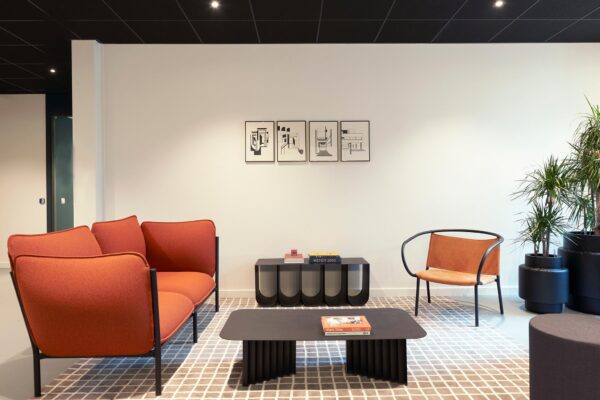 Modern ingerichte wachtruimte met een oranje bank, designstoel, zwarte salontafel, kunst aan de muur en grote kamerplanten.